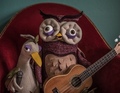 Eine Eulen-Puppe mit Gitarre und eine Vogel-Puppe mit Mikrofon sitzen auf einem roten Sessel vor grünem Hintergrund.