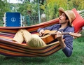 Mann mit Strohhut liegt in bunter Hängematte im Garten und spielt Gitarre. Im Hintergrund steht ein blauer Stuhl.