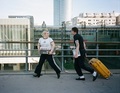 Zwei Personen gehen auf einer Brücke, eine trägt ein Keyboard, die andere zieht einen gelben Koffer. Gebäudekulisse im Hintergrund.