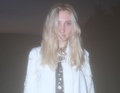 Person mit langen, blonden Haaren trägt eine weiße Jacke vor einem dunklen Hintergrund. Die Aufnahme wirkt verschwommen.