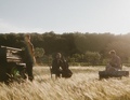 Drei Personen musizieren in einem sonnendurchfluteten Feld: eine am Klavier, eine mit einem Cello und eine auf einer Kiste sitzend.