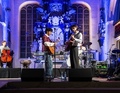 Musiker spielen auf einer Bühne in einer Kirche; zwei Gitarristen in der Mitte, links ein Cellist, hinten Schlagzeug.