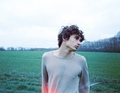 Ein Mann mit lockigem Haar in einem grauen Shirt steht auf einem Feld. Der Himmel ist bewölkt, und die Bäume im Hintergrund sind kahl.