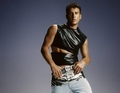 Ein Mann in einem schwarzen, asymmetrischen Oberteil und blauen Jeans posiert vor einem blauen Hintergrund.