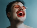 Eine Person lacht mit weit geöffnetem Mund, trägt eine Sonnenbrille und roten Lippenstift. Der Hintergrund ist blau.