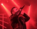 Eine Person singt leidenschaftlich in ein Mikrofon in einem roten, dramatisch beleuchteten Raum. Sie trägt eine Lederjacke.