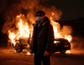 Eine vermummte Person steht vor einem brennenden Auto in der Nacht. Flammen und Rauch steigen in die Dunkelheit auf.
