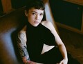 Eine Person mit einem schwarzen Kleid sitzt auf einem Stuhl. Ein bunter Arm-Tattoo ist sichtbar. Sie schaut direkt in die Kamera.