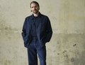 Ein Mann steht vor einer abgenutzten Wand, trägt eine dunkelblaue Jeansjacke und passende Jeans, die Hände in den Taschen.