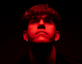 Eine Person mit Locken schaut nach oben. Das Gesicht ist rot beleuchtet und der Hintergrund ist dunkel.