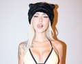 Eine Person mit langem blondem Haar trägt eine schwarze Mütze mit Katzenohren und ein gelbes Oberteil. Sie hat Tattoos auf dem Arm.