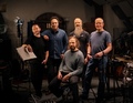 Fünf Männer in einem Musikstudio, einer sitzt mit Notenblatt, Schlagzeug im Hintergrund, alle lächeln in die Kamera.
