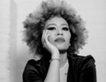 Person mit Afro, stützt den Kopf auf die Hand, trägt schwarzen Blazer und Kette, schaut ernst in die Kamera. Schwarz-weiß Foto.