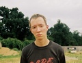 Junge steht ernst in einem offenen Gelände mit Bäumen im Hintergrund. Er trägt ein dunkles T-Shirt mit rotem Schriftzug.