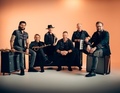Sechs Männer mit Musikinstrumenten, inklusive Gitarre, Saxophon und Keyboard, posieren lächelnd vor einem orangen Hintergrund.