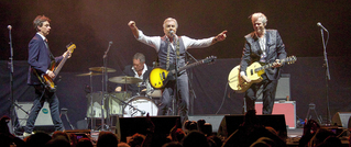 Vierköpfige Band spielt auf einer Bühne; Gitarristen rechts und links, Schlagzeuger hinten, Frontmann singt ins Mikrofon.