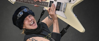 Eine Person mit schwarzem Fellhut und Sonnenbrille auf dem Kopf hält freudig eine weiße E-Gitarre hoch, vor grauem Hintergrund.