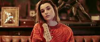 Eine Person in einem roten Pullover sitzt auf einem Sofa, hält eine Gitarre. Der Hintergrund zeigt eine Skulptur und gerahmte Bilder.