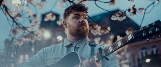 Ein Mann in einer Jeansjacke spielt Gitarre unter blühenden Baumzweigen. Der Hintergrund zeigt verschwommene Gebäude bei Dämmerung.
