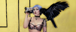 Eine Person mit blauem Bob hält eine Kamera ans Auge. Sie trägt Dessous. Ein großer schwarzer Vogel sitzt auf ihrer Schulter.