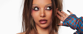 Person mit dramatischem Make-up, vollem Haar und markanten Nägeln, trägt bunt kariertes Kleidungsstück, schaut zur Seite.