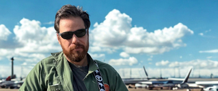 Ein Mann mit Sonnenbrille und grünem Hemd steht vor einem Flughafen mit geparkten Flugzeugen, blauer Himmel und Wolken im Hintergrund.