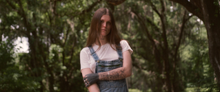 Eine Person mit langen Haaren steht in einem Wald, trägt ein weißes Shirt und eine blaue Latzhose. Ihr Arm ist tätowiert.