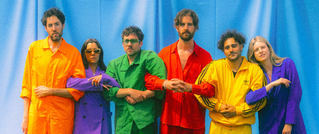 Sechs Personen in bunten Outfits (orange, lila, grün, rot, gelb) stehen Arm in Arm vor einem blauen Hintergrund.
