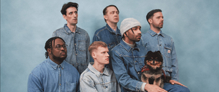 Sechs Personen in Jeanskleidung posieren vor einem blauen Hintergrund. Einer hält einen kleinen Hund auf dem Schoß.
