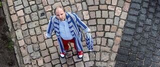 Ein Mann mit gestreiftem Bademantel und roten Hosen steht auf Kopfsteinpflaster und schaut nach oben. Er hält ein Handtuch.