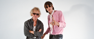 Zwei Männer mit Sonnenbrillen posieren vor einem weißen Hintergrund. Einer trägt ein graues Hemd, der andere eine pinke Jacke.