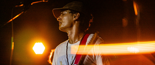 Ein Musiker mit Hut spielt Gitarre auf einer Bühne, beleuchtet von warmem Scheinwerferlicht.