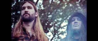 Zwei Personen mit langen Haaren stehen im Freien; der linke trägt einen Bart, der rechte eine Kapuze. Bäume im Hintergrund.