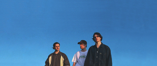 Drei Personen stehen vor einem klaren blauen Himmel, tragen Freizeitkleidung und blicken in verschiedene Richtungen.