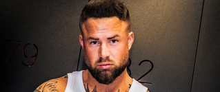 Ein Mann mit Bart und Tattoos trägt ein weißes Tanktop und schaut ernst. Der Hintergrund ist dunkel mit roten Zahlen.