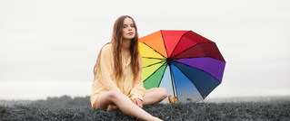 Frau in gelbem Kleid sitzt auf grauem Gras neben einem aufgespannten, regenbogenfarbenen Regenschirm; bewölkter Himmel.