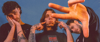 Drei junge Männer mit Tattoos posieren vor einem blauen Hintergrund, einer streckt die Hand in die Kamera, alle wirken entspannt.