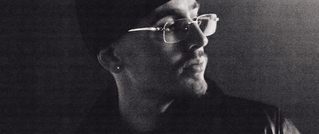 Ein Mann mit Brille und Mütze blickt nach rechts; schwarz-weiß Aufnahme mit starker Schattenwirkung.