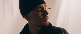 Ein Mann mit Brille und Bart trägt eine schwarze Mütze und Jacke, schaut nach rechts. Der Hintergrund ist weich und hell.
