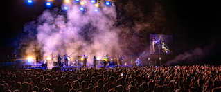 Eine Menschenmenge bei einem nächtlichen Konzert, Bühne erleuchtet, Künstler performen, Rauch im Hintergrund, Videoleinwand rechts.
