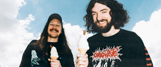 Zwei lächelnde Männer mit langen Haaren halten Softeis in der Hand, tragen schwarze Shirts mit Bandmotiven, blauer Himmel im Hintergrund.