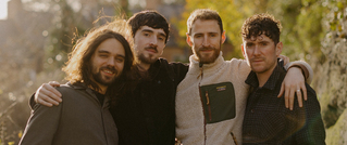 Vier Männer stehen im Freien nebeneinander, legen die Arme umeinander und lächeln. Im Hintergrund sind Bäume und Gebäude.