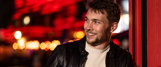 Ein Mann in einer schwarzen Lederjacke lächelt nach links. Der Hintergrund ist unscharf mit roten Lichtern und Bokeh-Effekten.