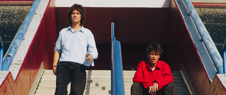 Zwei Personen auf einer Treppe: eine steht links mit weißem Hemd, die andere sitzt rechts in rotem Pullover, beide vor blauem Geländer.