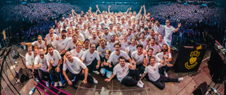 Eine große Gruppe von Menschen in weißen T-Shirts posiert lächelnd auf einer Bühne vor einem vollen Publikum in einer Arena.