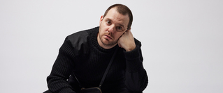 Eine Person in schwarzem Pullover sitzt vor einem neutralen Hintergrund, stützt ihren Kopf mit der Hand ab und blickt nachdenklich.