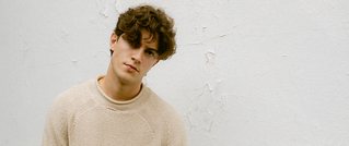 Ein Mann mit lockigem Haar trägt einen beigefarbenen Pullover und steht vor einer leicht abblätternden weißen Wand.