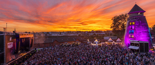 Ein Open-Air-Konzert bei Sonnenuntergang. Große Menschenmenge vor einer Bühne links und ein beleuchteter Turm rechts.
