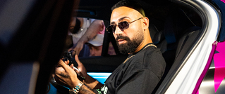 Ein Mann mit Bart und Sonnenbrille sitzt in einem Auto und hält etwas in der Hand. Er trägt ein dunkles Shirt.