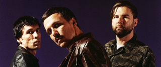 Drei Männer vor einem violetten Hintergrund. Sie tragen Leder- und Camouflagejacken und blicken ernst in die Kamera.